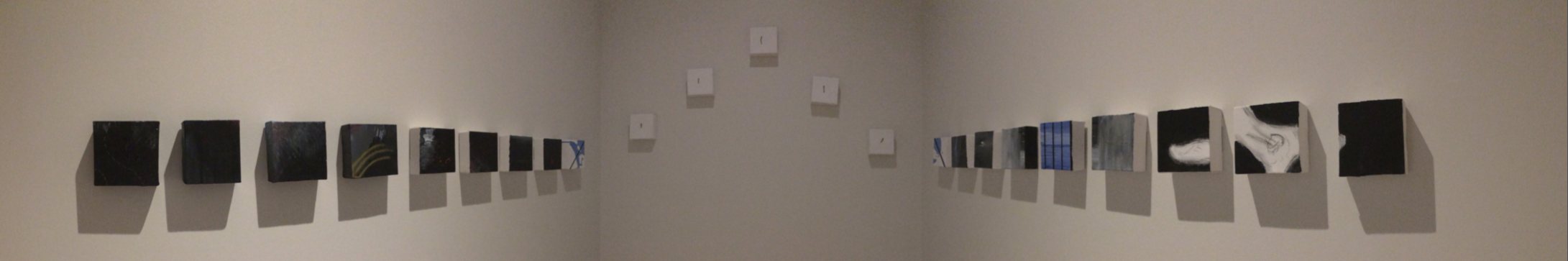 Twenty-three small square canvases hung in a line around a gallery room, mostly dark, five white on the far wall.