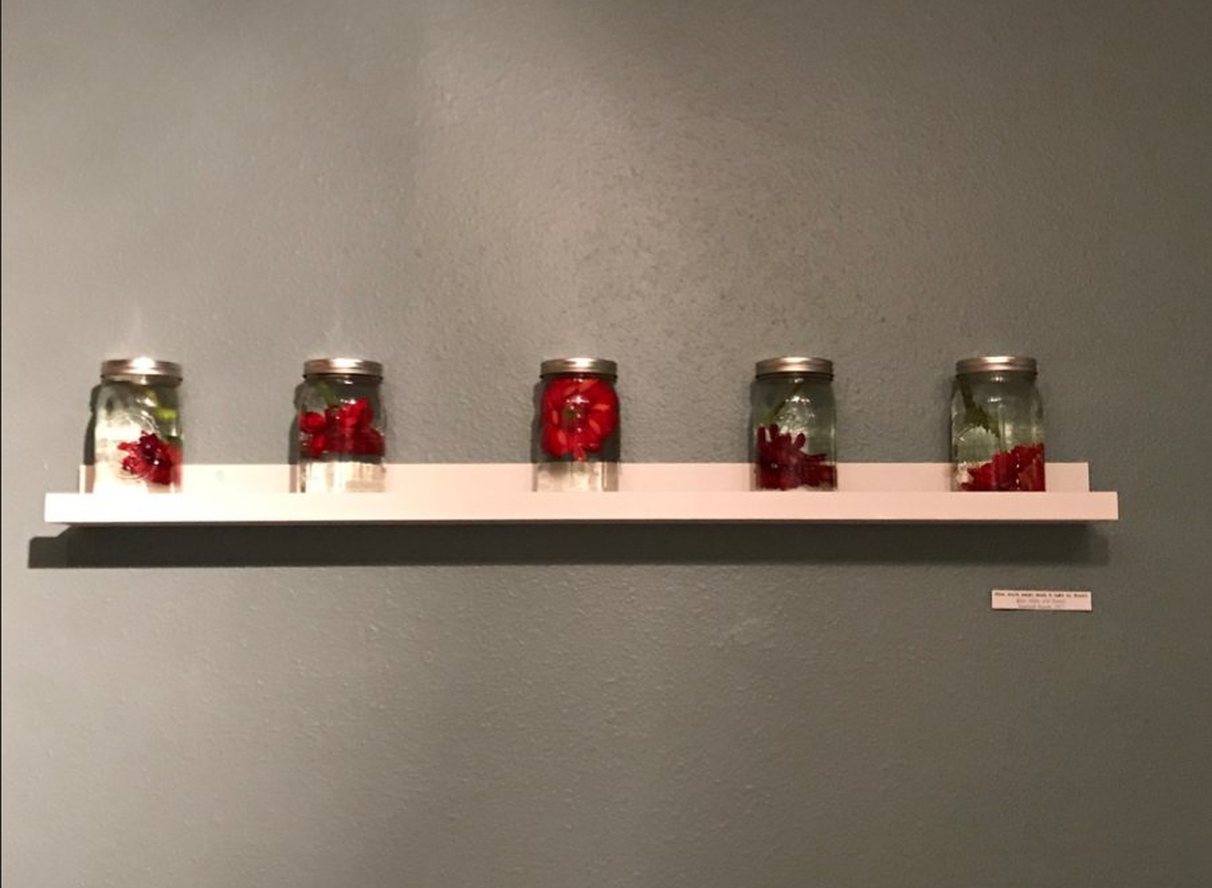 Five mason jars of clear water and red flowers on a white shelf against a grey-green wall.